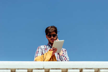 Handsome young man walking and talking on mobile phone in the city and looking in tabletの写真素材
