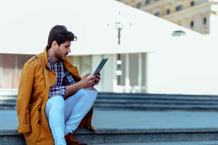 Young handsome manager in yellow coat work on tablet while sitting in downtown