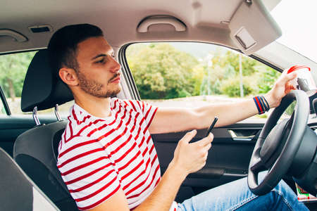 Young man is looking forward while is sitting in his car and is having coffee in one hand and his phone in the otherの写真素材
