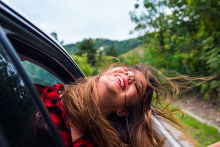 A girl is with her head out of a car window while is driving in nature, smiling and having fun
