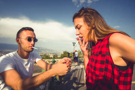 Guy is lightening a cigarette to the girl outdoors, spending time together on a sunny dayの写真素材
