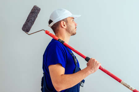Side view of guy in blue uniform while hold paint roller on white wallの写真素材