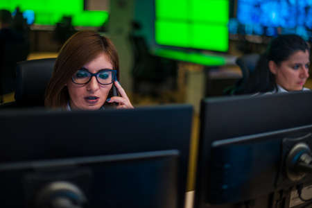Female security guard sitting and monitoring modern CCTV cameras in surveillance room, talking on a cellphone.の写真素材