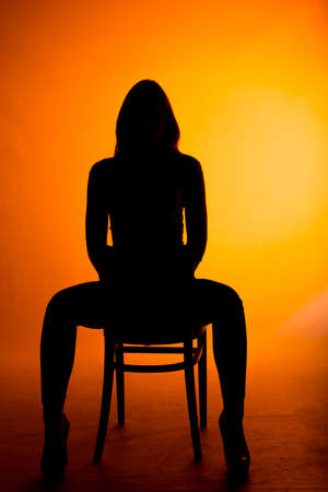 Body shot of a cool modern silhouette girl sitting on cheir posing in black top and blue jeans isolated on orange wallの写真素材
