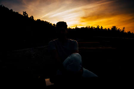 Relaxed african american young man sitting and waiting on bench against sunset sky