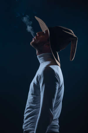 Portrait of a handsome young man with cowboy hat smoking cigarette in a studio on high contrast and dark backgroundの写真素材