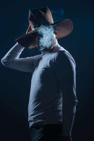 Portrait of a handsome young man with cowboy hat smoking cigarette in a studio on high contrast and dark backgroundの写真素材