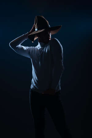 Portrait of a sexy young man with cowboy hat posing in a studio on high contrast and dark backgroundの写真素材