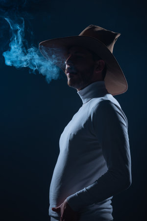 Portrait of a handsome young man with cowboy hat smoking cigarette in a studio on high contrast and dark backgroundの写真素材