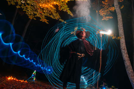 Sorcerer (magician) casting a spell while standing in the magical woods. Holding his magical staff. Magic illumination. Autumn holiday celebration. Mystery and nightmare conceptの写真素材