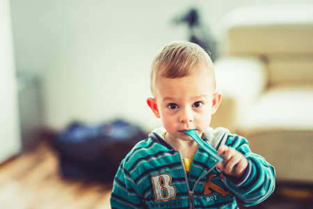 A little baby boy is standing in the living room with a spoon in his mouthの写真素材