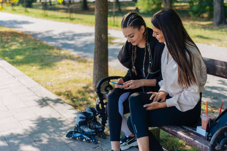 Two young girls are sitting on a bench outdoor while one of them is showing something to her friend on the mobile phoneの写真素材