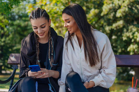 Two young girls are sitting on a bench outdoor while one of them is showing something to her friend on the mobile phoneの写真素材