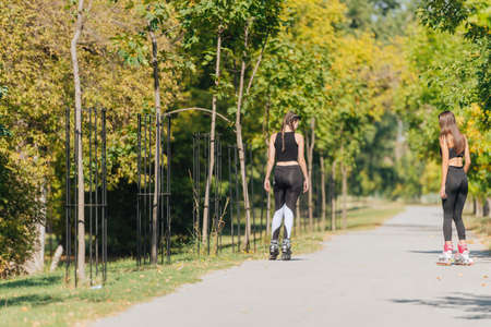 Rearview of two young fit women on roller skates riding outdoors on urban street in the park.の写真素材
