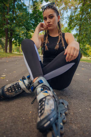 Astonishing young woman with hair braids posing in  roller skates while sitting on a road in the parkの写真素材
