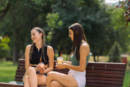 Close-up of two young girlfriends sitting on a bench in summer park chatting and having funの写真素材