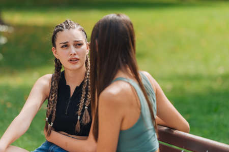 Two girls sitting on a bench in a park and talking to each other on a sunny day.