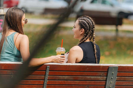 Two girls sitting on a bench in a park and talking to each other on a sunny day.の写真素材