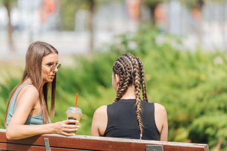 Two girls sitting on a bench in a park and talking to each other on a sunny day.の写真素材