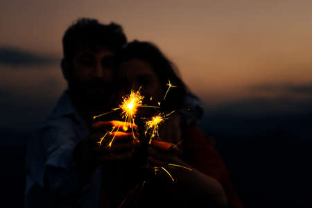 Party, event and holiday concept - man and woman fooling around with sparklers against sunset sky