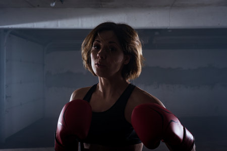 Silhouette portrait of young attractive fitness woman exercising punching with red boxing gloves in a garageの写真素材