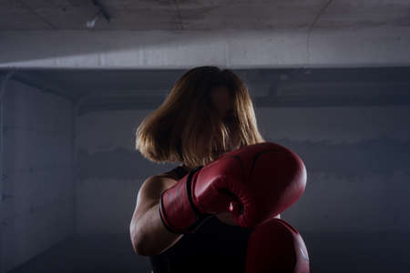 Silhouette portrait of young attractive fitness woman exercising punching with red boxing gloves in a garageの写真素材