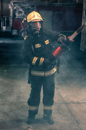 Portrait of a brave firefighter standing confident wearing full protective equipment, turnouts and helmet. Dark background with smoke and blue light.の写真素材