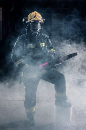 Portrait of a brave firefighter standing confident wearing full protective equipment, turnouts and helmet. Dark background with smoke and blue light.の写真素材