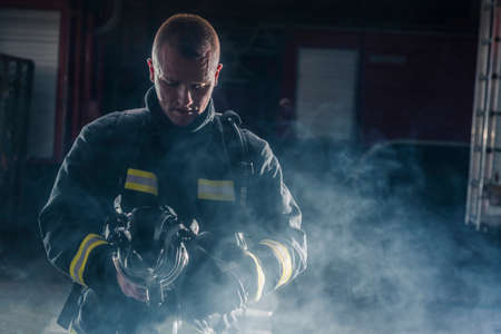 Portrait of a fireman wearing firefighter turnouts and helmet. Dark background with smoke and blue...