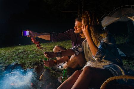 Young couple making selfie, drinking beer and baking sausages on the campfire in the forest hill in the night. Relaxing romantic get-away weekendの写真素材