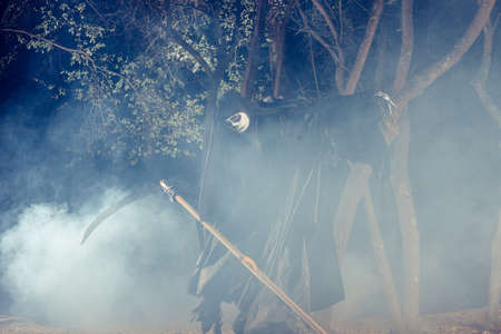 Portrait of grim reaper, the angel of death ( Azrael ) lurking through the woods at night surrounded with mist, smoke in a mystical forest holding his shythe.の写真素材