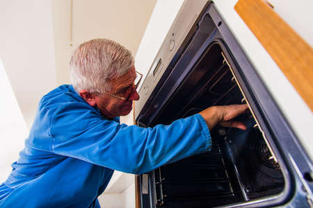 Mature handyman in blue working suit fixing the oven in the kitchen