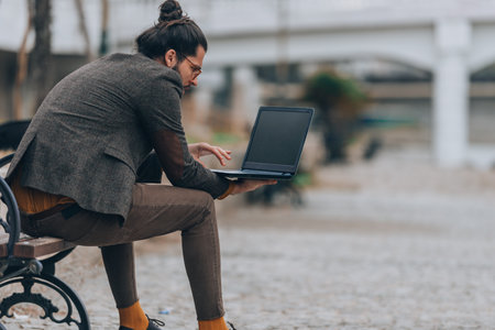 Handsome man with a long beard sitting on a bench near a river and using pcの写真素材