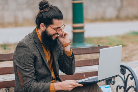 Fashionable male wearing stylish modern clothing talking on a phone by the river while looking in laptopの写真素材