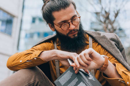 Handsome man with a long beard in stylish clothes checking his watch at the city centerの写真素材