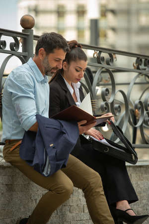 Two urban business people are smiling and talking about the company while sitting on the wall