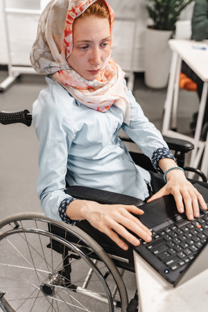 Female paralyzed employee in wheelchair working on lap top. Muslim disabled employee in wheelchair wearing hijab working on lap topの写真素材