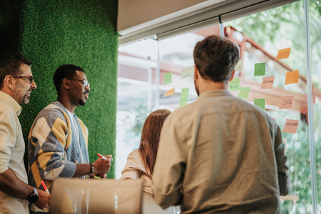 Diverse colleagues brainstorm, discuss and plan projects. They write ideas on sticky notes over a...
