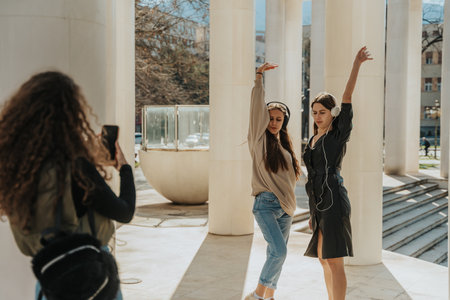 Lovely brunette girls dancing with one hand up while listening to music on their headphones. Their curly haired friend is taking a photo of themの写真素材