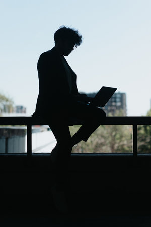Silhouette photo of businessman sitting and using lap top placed on a metal fenceの写真素材