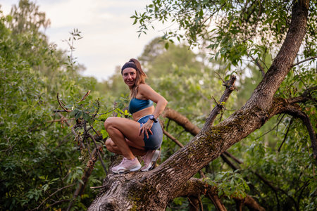 Fit girl exercising outdoors in a green park, climbing tree. Demonstrates flexibility, strength, and healthy lifestyle.の写真素材