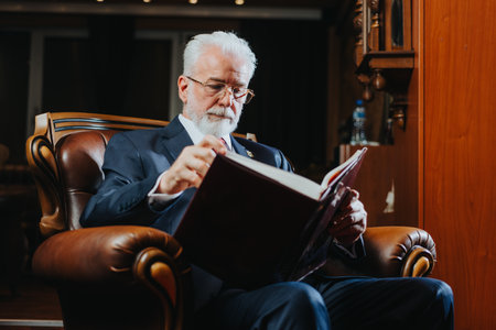 Elderly college rector sitting in modern armchair reading a book or watching old pictures in album, back to memories.の写真素材