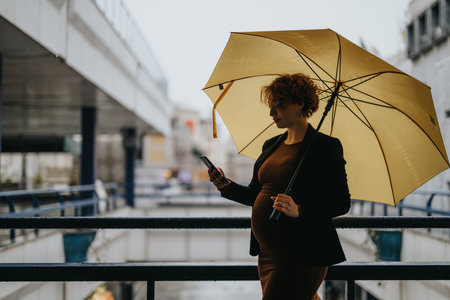 Pregnant businesswoman chatting on phone while holding umbrella and standing at big building center.の写真素材