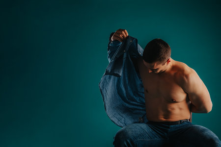 Fit male gym instructor putting shirt on while sitting on a chair and posing on turquoise background in studio.の写真素材