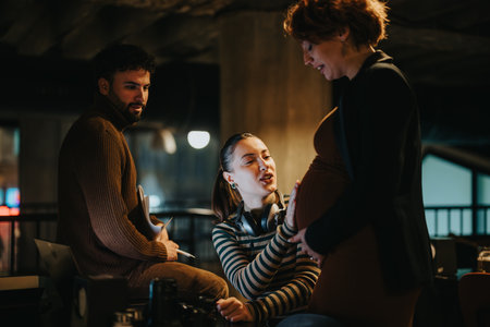 Stunning woman gently touching her pregnant colleagues stomach while working together at the office. They are having a conversation and smiling.の写真素材