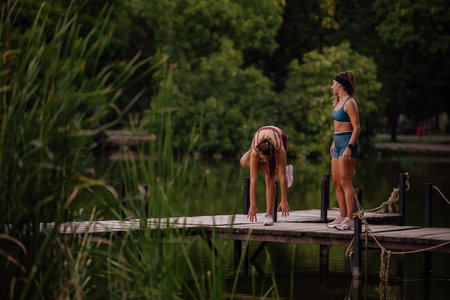 Fit women warm up together on a park bridge, stretching and preparing for a workout in a green outdoor setting.の写真素材