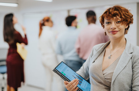 Ginger businesswoman smiling and posing with lap top at the office while her colleagues brainstorming in the back, Copy space.の写真素材