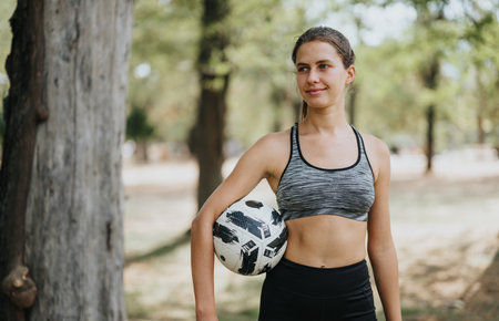 Fit woman posing in a park with a soccer ball after training, enjoying the sunny day and nature.の写真素材
