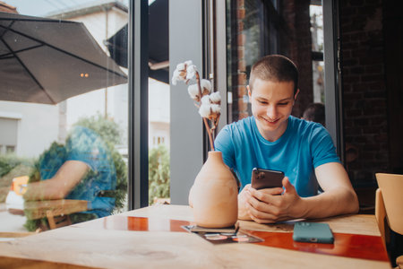 A school teenager enjoys a coffee alone at a cafe bar, browsing social medias and messaging on his smart phone after school.の写真素材