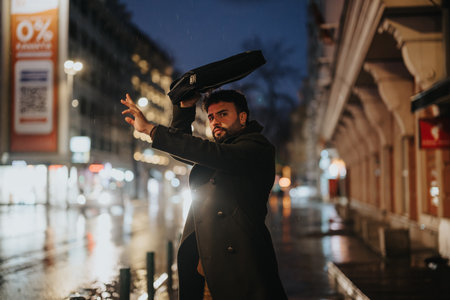 A stylish businessman stands in a busy city, covering his head from the rain with lap top case. He is waiting for a bus to go home after work.の写真素材